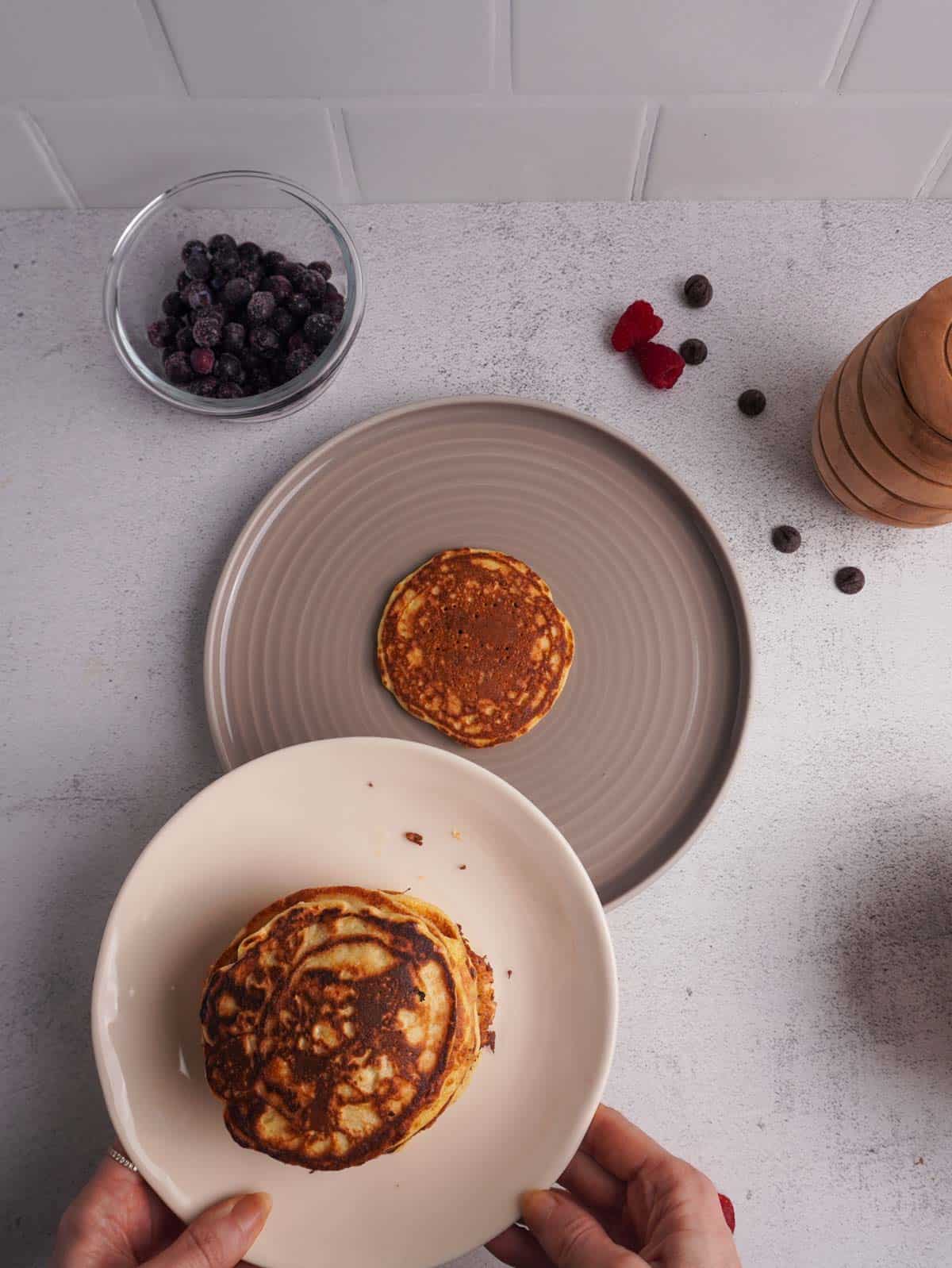 A pancake on a plate with another plate of more pancakes ready to be added to the stack.