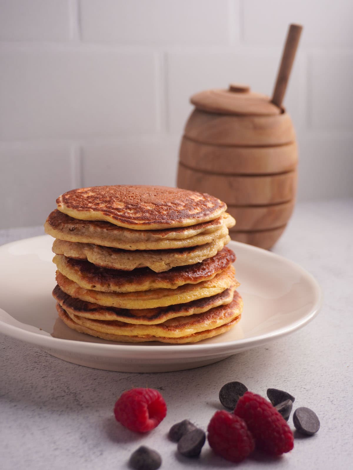 A plate pf pancakes stacked high with a honey jar in the background and berries in the foreground.