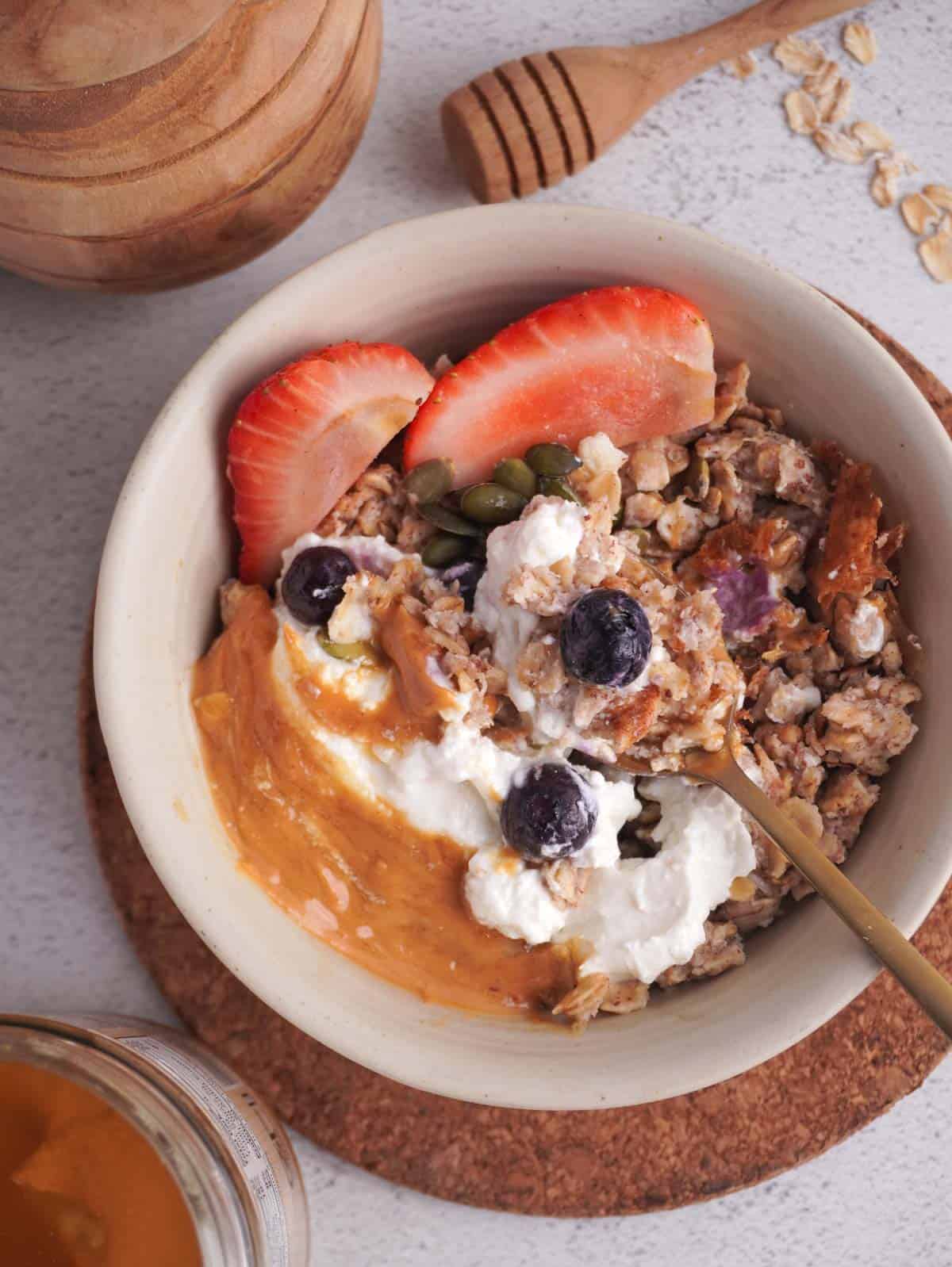 A bowl of scrambled oats with greek yogurt, peanut butter and berries on top, with a spoon in the middle taking a spoonful out of the bowl.